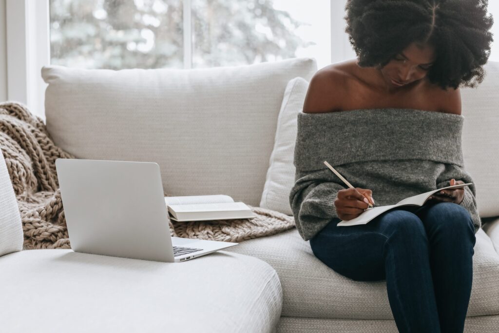 Black woman taking notes while sitting on a white couch with laptop beside her