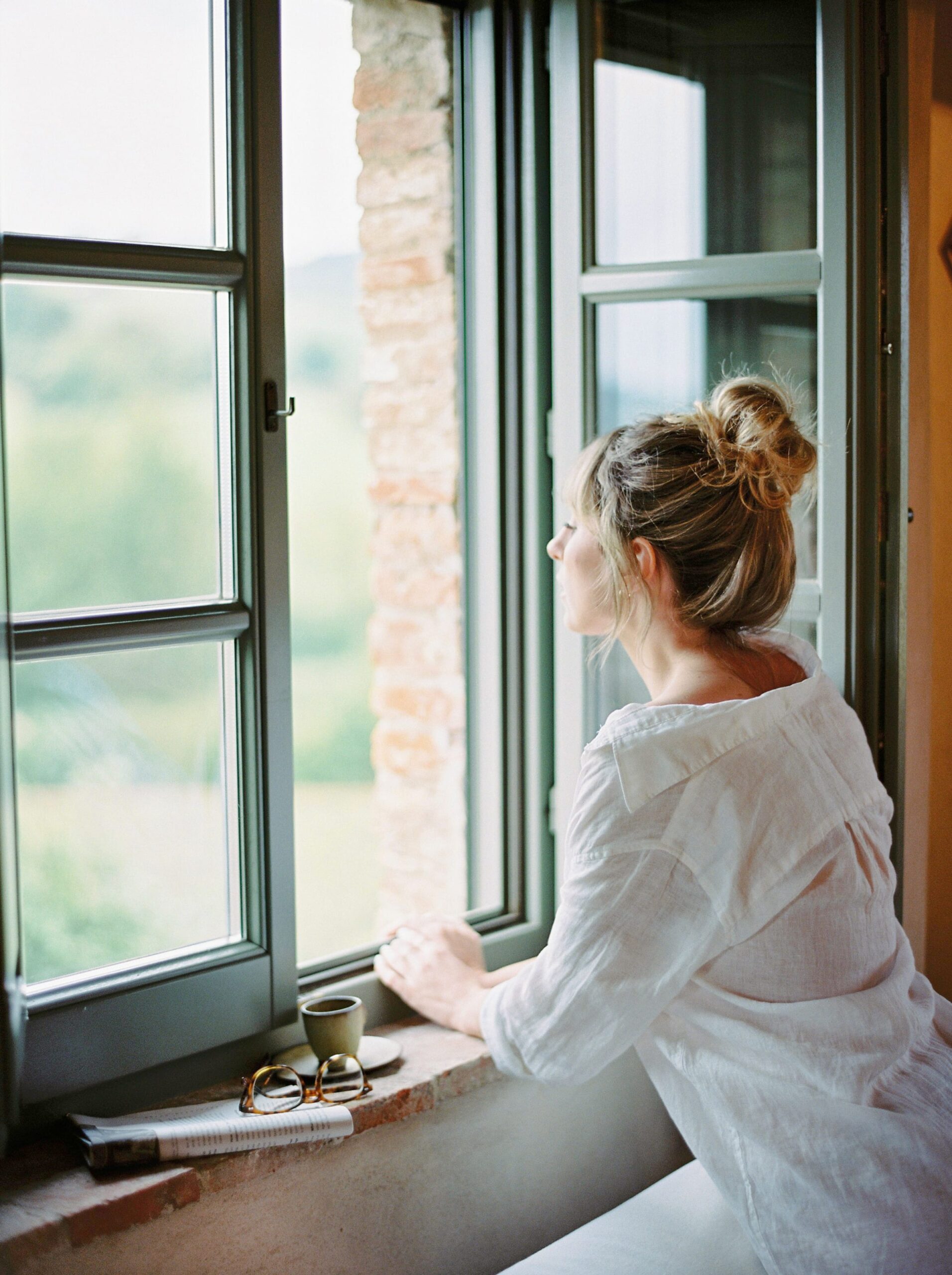 woman facing away looking out window with hair up in a bun wearing a white shirt