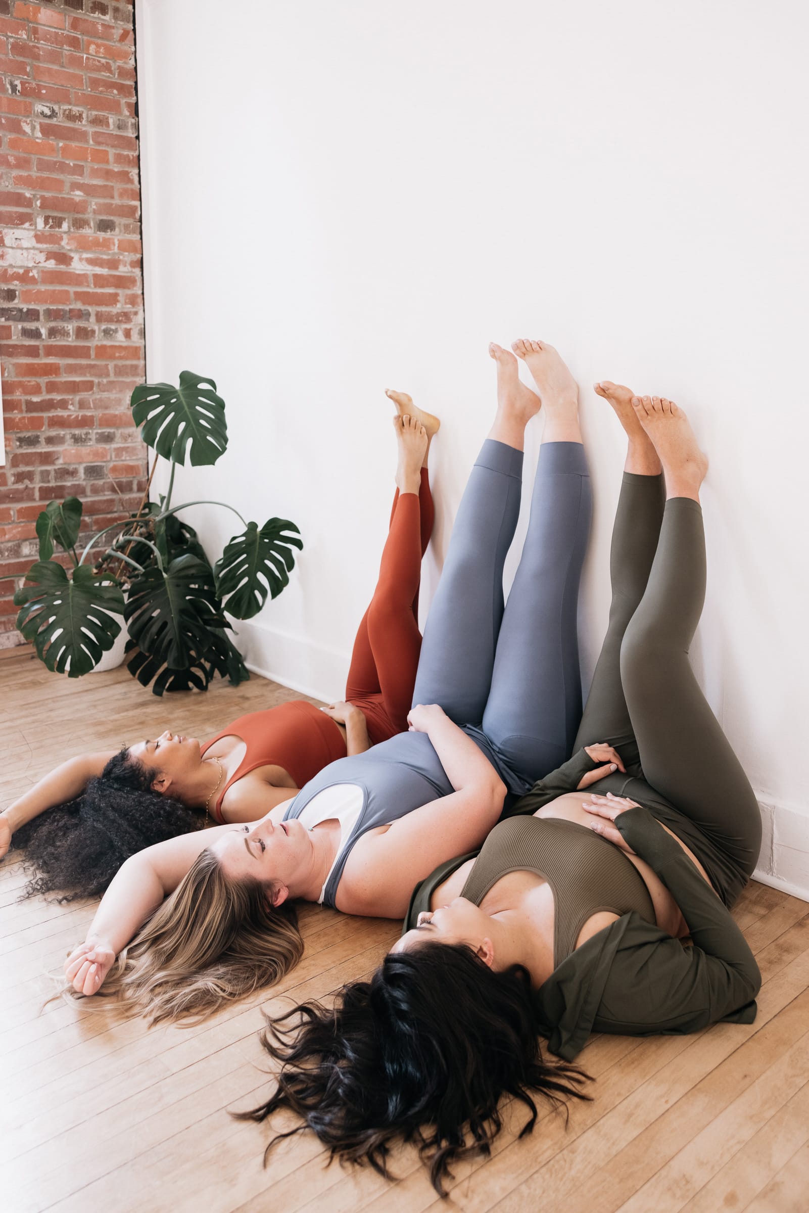 Three female presenting people wearing gym kit lie on their backs with their legs up against a white wall.