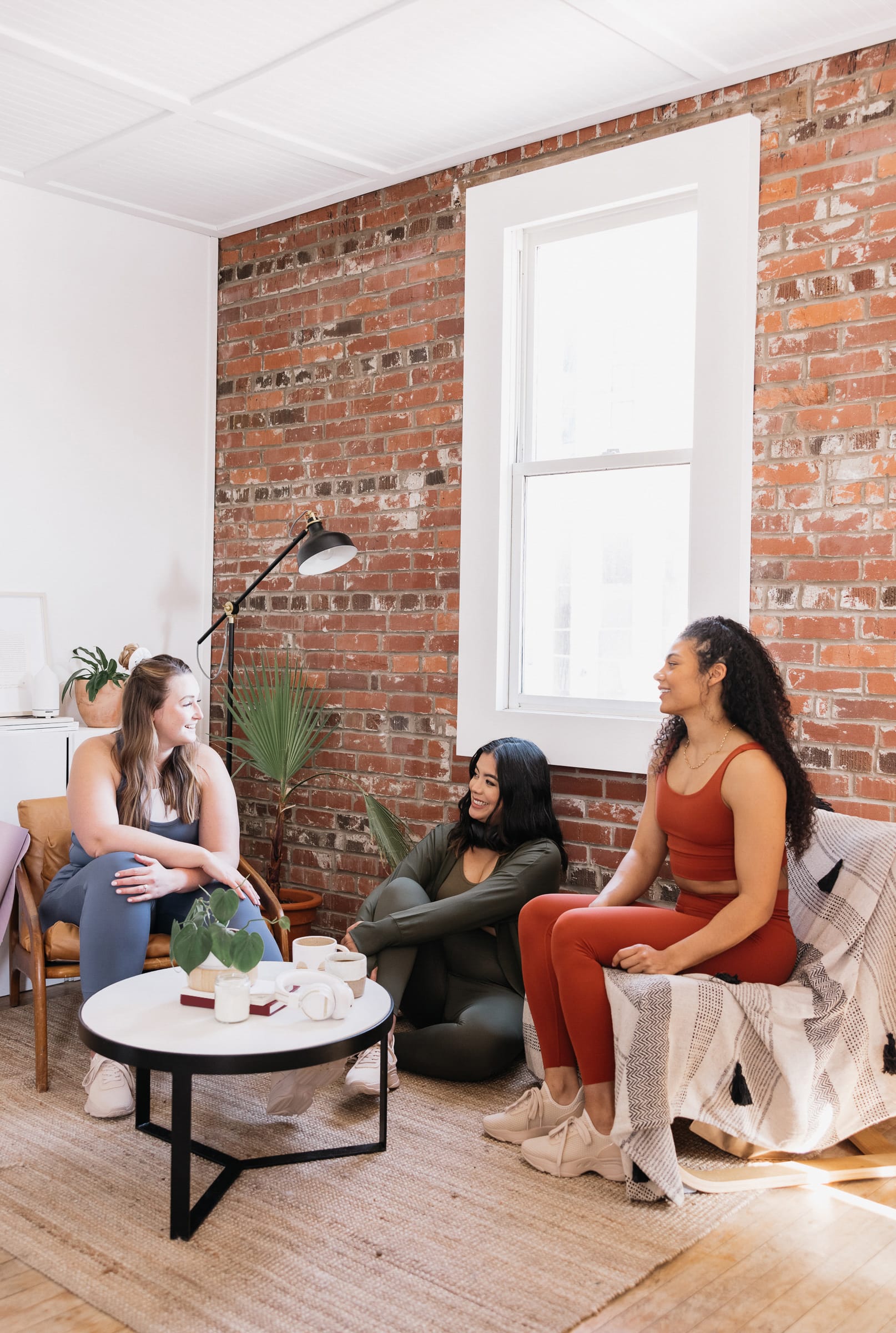 Three female presenting friends sit in varying positions and chat in front of a brick wall with a window in it