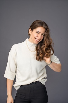 Hypermobility specialist osteopath Alex stands facing camera wearing a white sweater with her brown hair down. She is smiling.