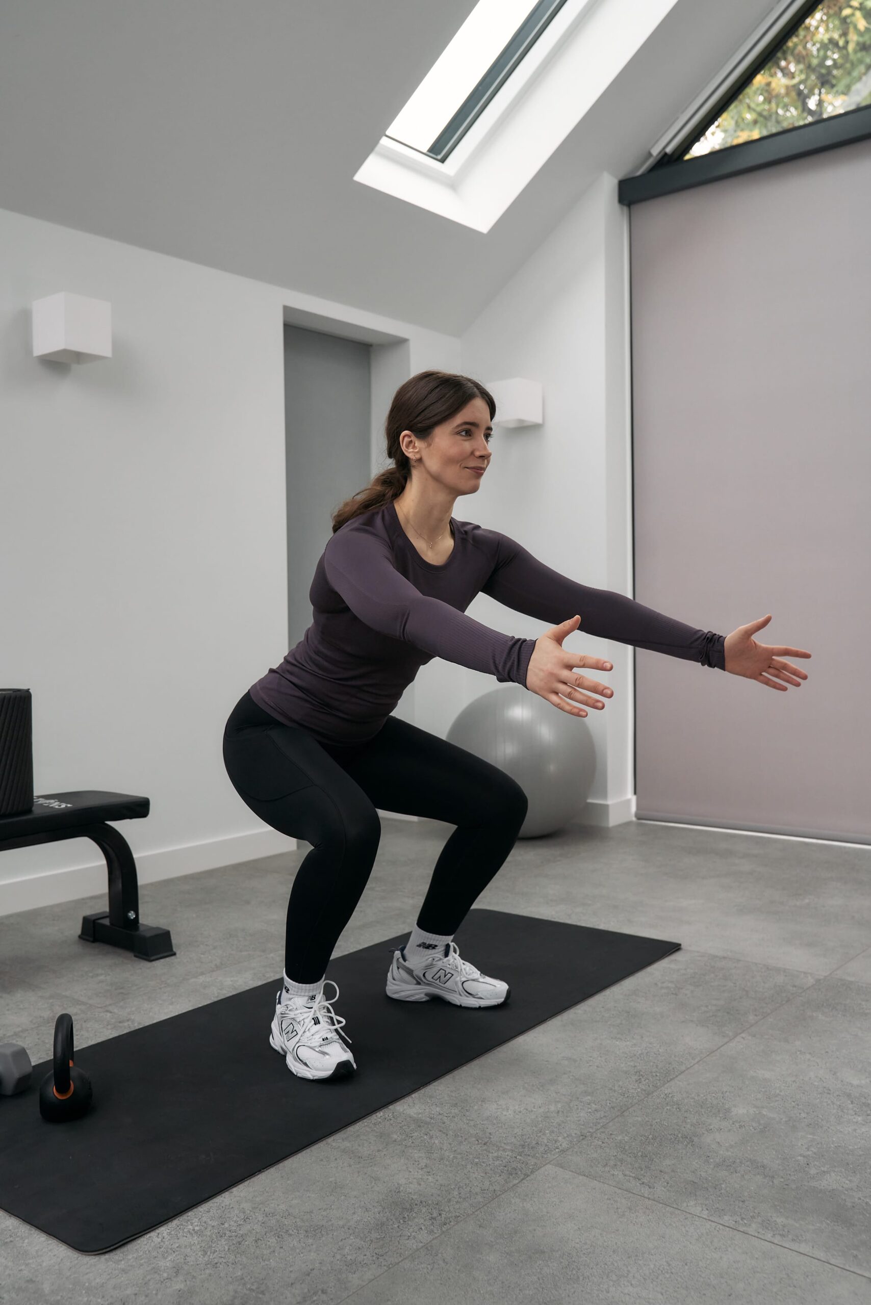 Female hypermobility expert wearing a purple top, black yoga pants, and white shoes demonstrates how to do a squat while smiling. She stands on a black yoga mat in a room with white walls and a grey floor.