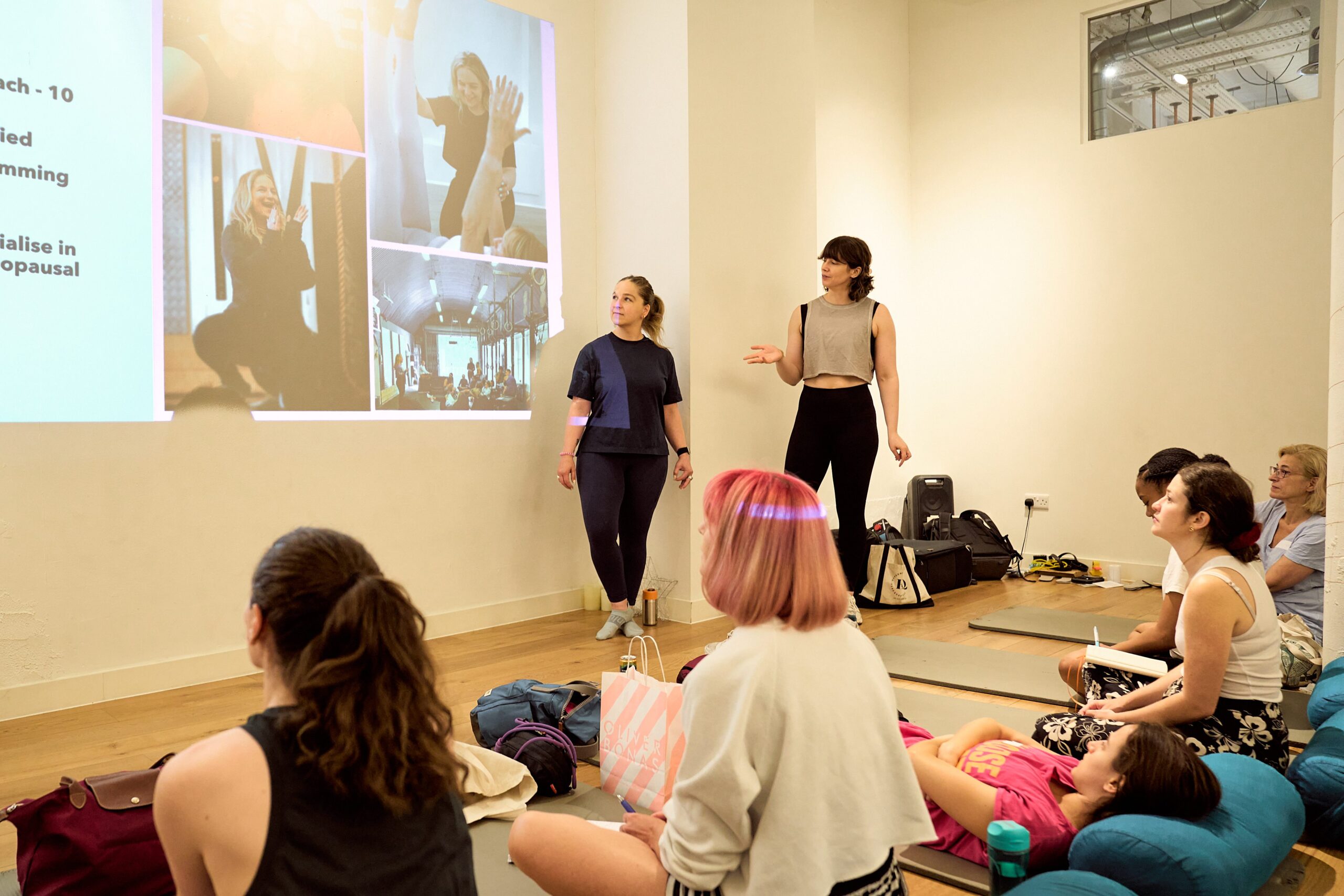Alex and Karima stand in front of an audience during the Hypermobility Strength Training workshop with a slide presentation showing images of Karima lifting weights on the wall behind them.
