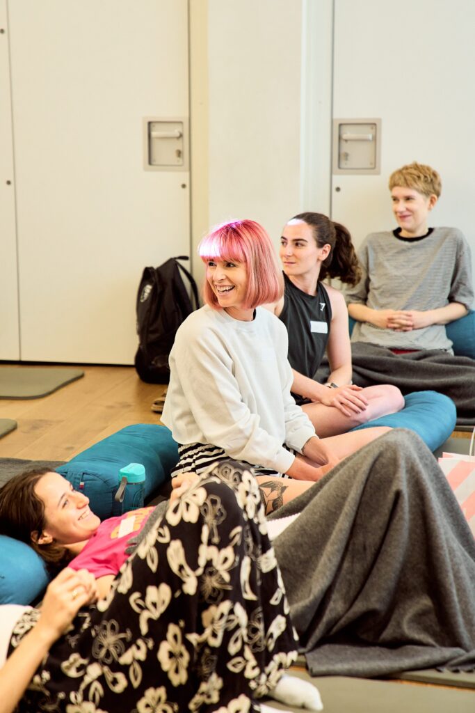 Workshop attendees sit on the floor during the Hypermobility Strength Training workshop. A woman with pink hair is seated in the centre wearing a white shirt. 